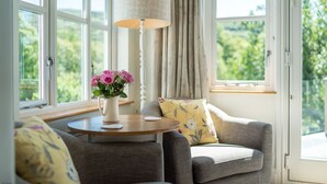 Una habitación acogedora con un sillón gris, una mesita redonda con una jarra blanca y flores rosadas, y una lámpara de pie alta.