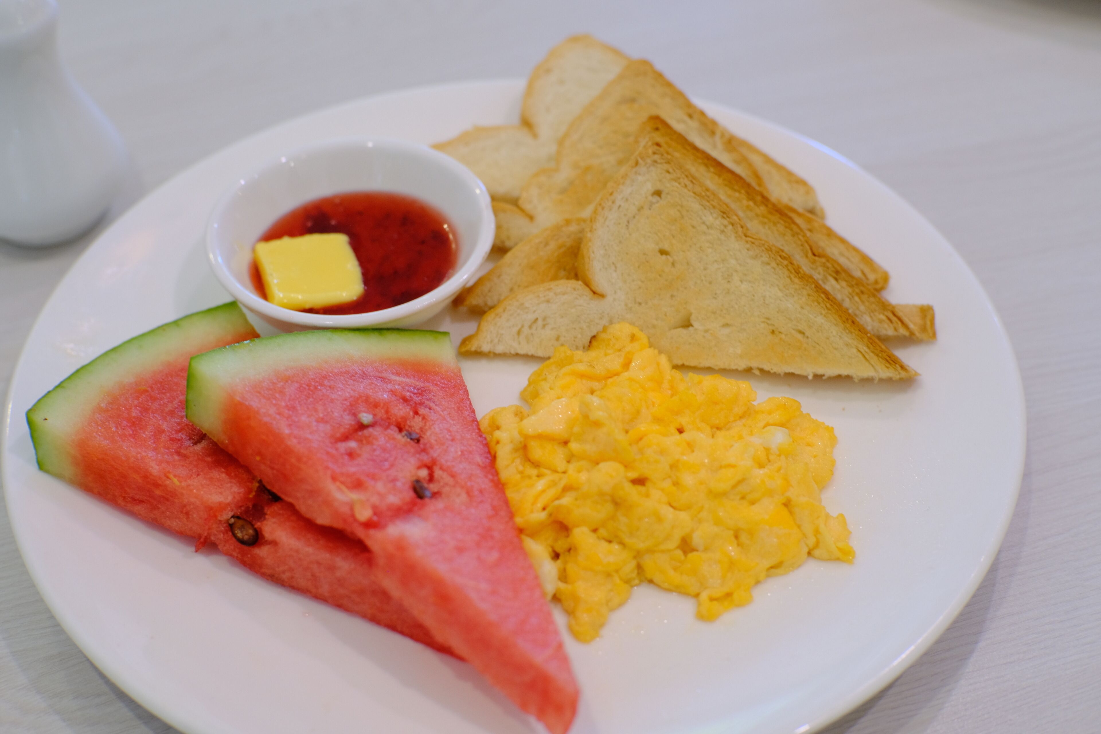 A plate with toast, scrambled eggs, watermelon, and a small bowl of jam.