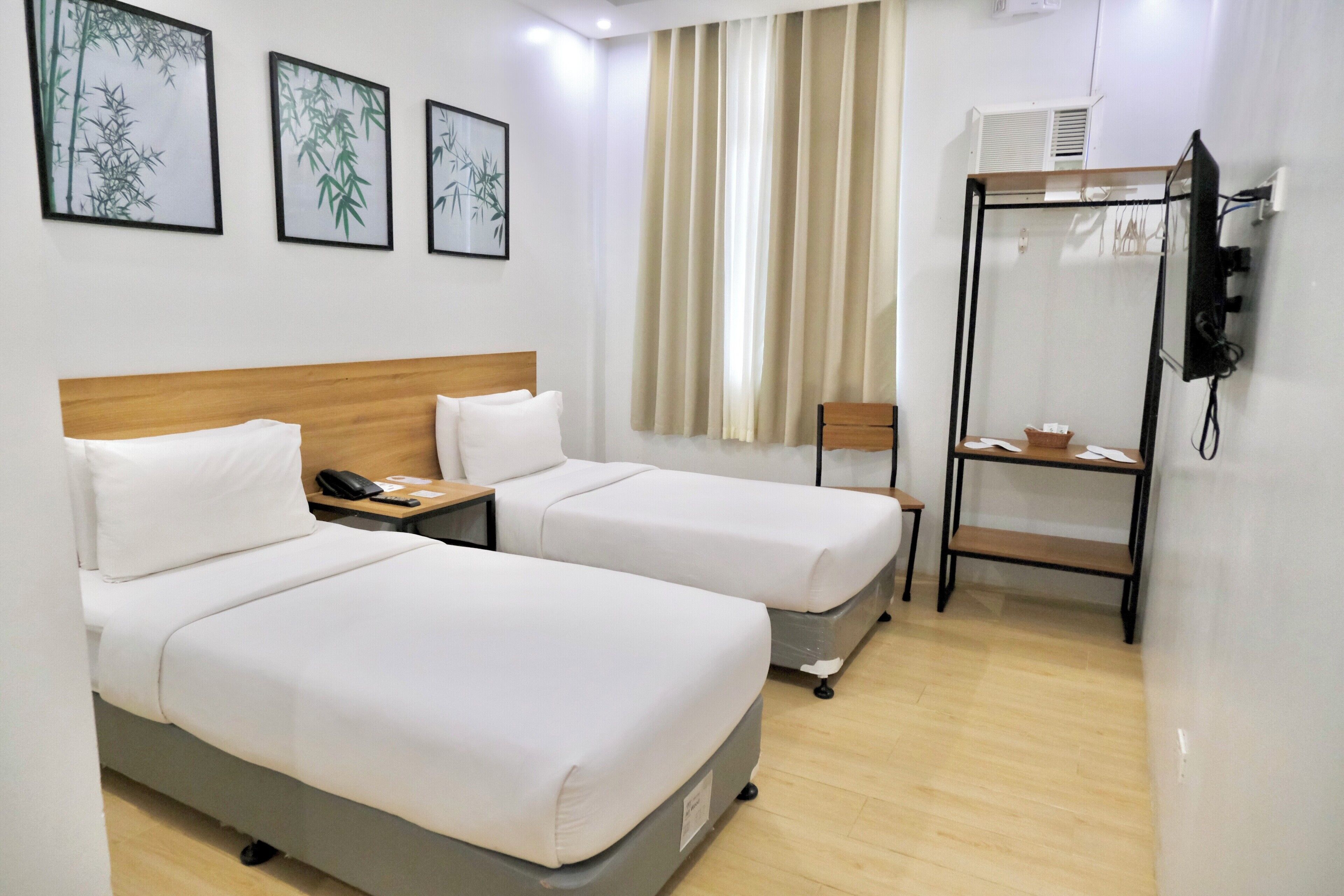 A hotel room with two beds, a wooden headboard, a desk with a phone, a chair, a shelf with items, and a window with curtains.