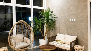 A room with a large window, a wicker chair, a potted plant, a sofa, and a small round table. - Bambu Suites (Puerto Princesa)