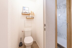 Un baño pequeño con inodoro, un estante con un cuadro enmarcado y una zona de ducha con azulejos de mármol.