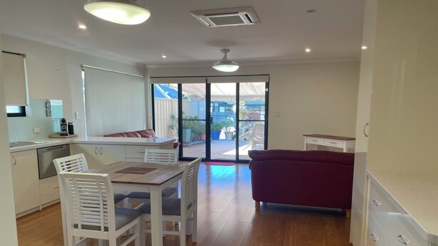 A modern kitchen with white cabinets, a dining area with a wooden table and chairs, and a sliding glass door leading to an outdoor patio. - Fully Self contained 1bedroom spacious unit adjoining rear of owners home. (Jandakot)