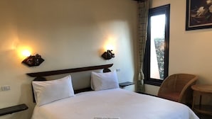 Una cama bien hecha con ropa blanca, dos lámparas fijadas a la pared, una silla de mimbre y una ventana con cortinas.