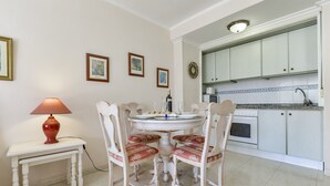 A dining area with a round table and four chairs, a small side table with a lamp, and a kitchen with white cabinets and a tiled backsplash.