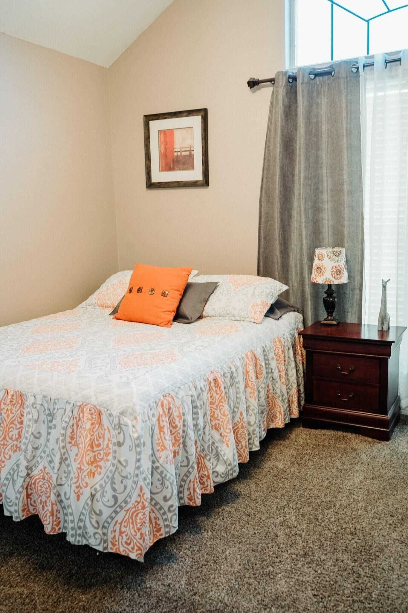 A bedroom with a bed, a nightstand, a lamp, a framed picture on the wall, and a window with curtains.