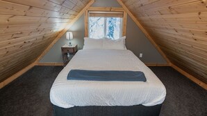 Une chambre avec un plafond en pente, un lit avec une literie blanche, une table de chevet et une fenêtre donnant sur des arbres.