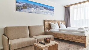 A modern room with a sofa, a wooden coffee table with a bowl of fruit, a bed with white bedding, and a large window with sheer curtains.