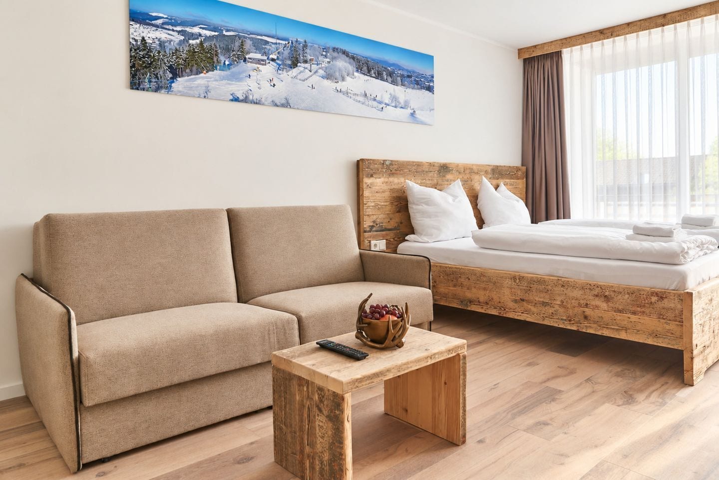 A modern room with a sofa, a bed with a wooden headboard, a small wooden table with a bowl of fruit, and a painting of a snowy landscape on the wall.