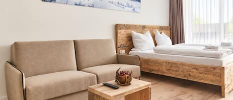 A modern room with a sofa, a bed with a wooden headboard, a small wooden table with a bowl of fruit, and a painting of a snowy landscape on the wall.