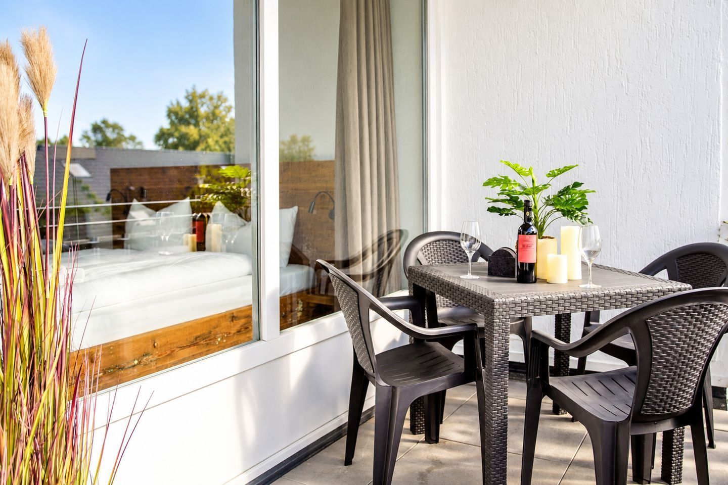 A modern outdoor dining area with wicker furniture, a glass table, and a view of a bedroom through a large window.