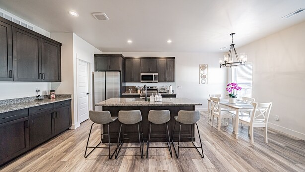 A modern kitchen with dark cabinets, a central island with bar stools, a dining area with a white table and chairs, and a chandelier.