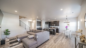 A modern living room with a sectional sofa, a dining area with a white table and chairs, and a kitchen with dark cabinets and stainless steel appliances.