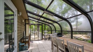 A covered patio with a table and chairs, a small table with a chair, and a view of a wooded area through large windows.