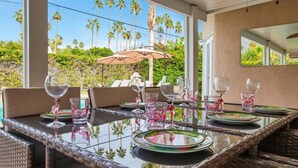 A dining table set with glassware and plates, surrounded by wicker chairs, with a view of palm trees and a large umbrella. - Tahquitz River Estates Oasis | Walled In Pool+Spa (Palm Springs)