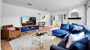 A modern living room with a blue sofa, a brown leather couch, and a glass coffee table. There is a flat-screen TV mounted on the wall, a fireplace, and a dining area in the background. - Tahquitz River Estates Oasis | Walled In Pool+Spa (Palm Springs)