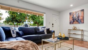 A modern living room with a dark blue sofa, a glass coffee table, and a view of trees through a large window. - Tahquitz River Estates Oasis | Walled In Pool+Spa (Palm Springs)