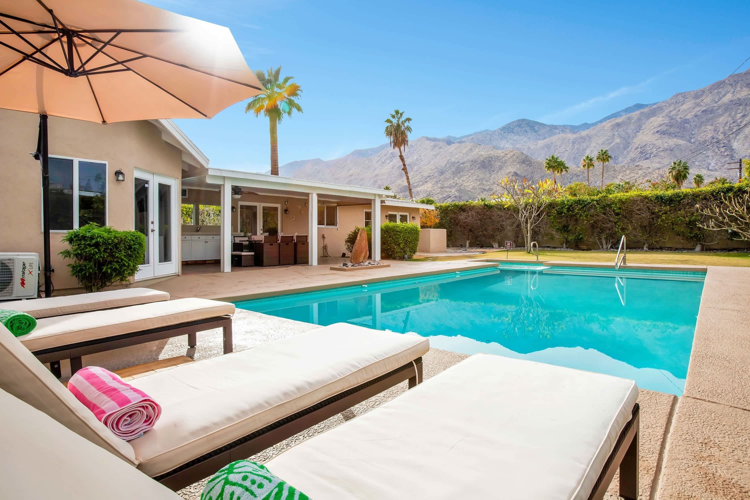 A pool area with lounge chairs and an umbrella, surrounded by mountains and palm trees.