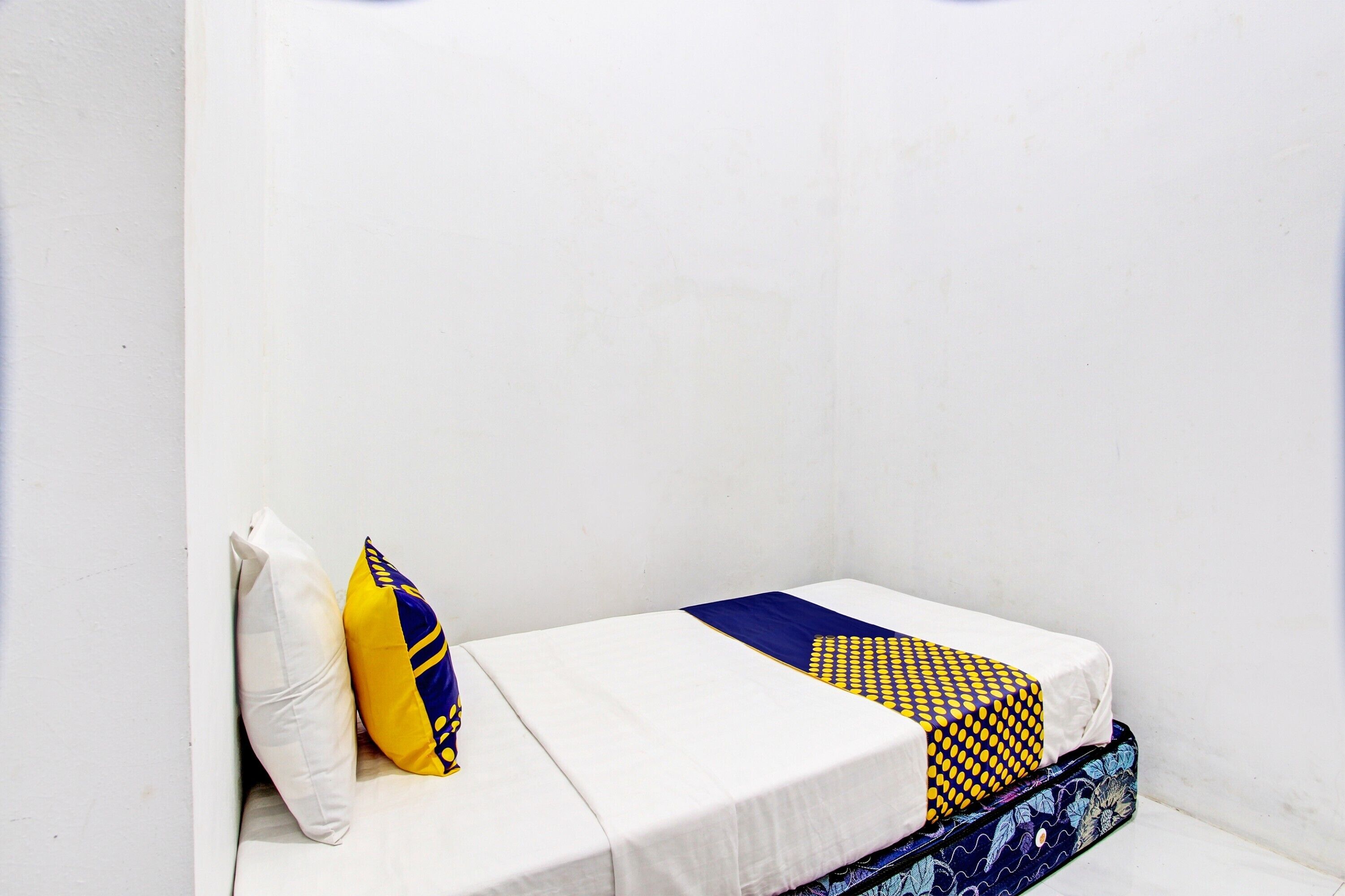 A small, single-bed room with a white mattress, a blue and yellow patterned blanket, and a white pillow.