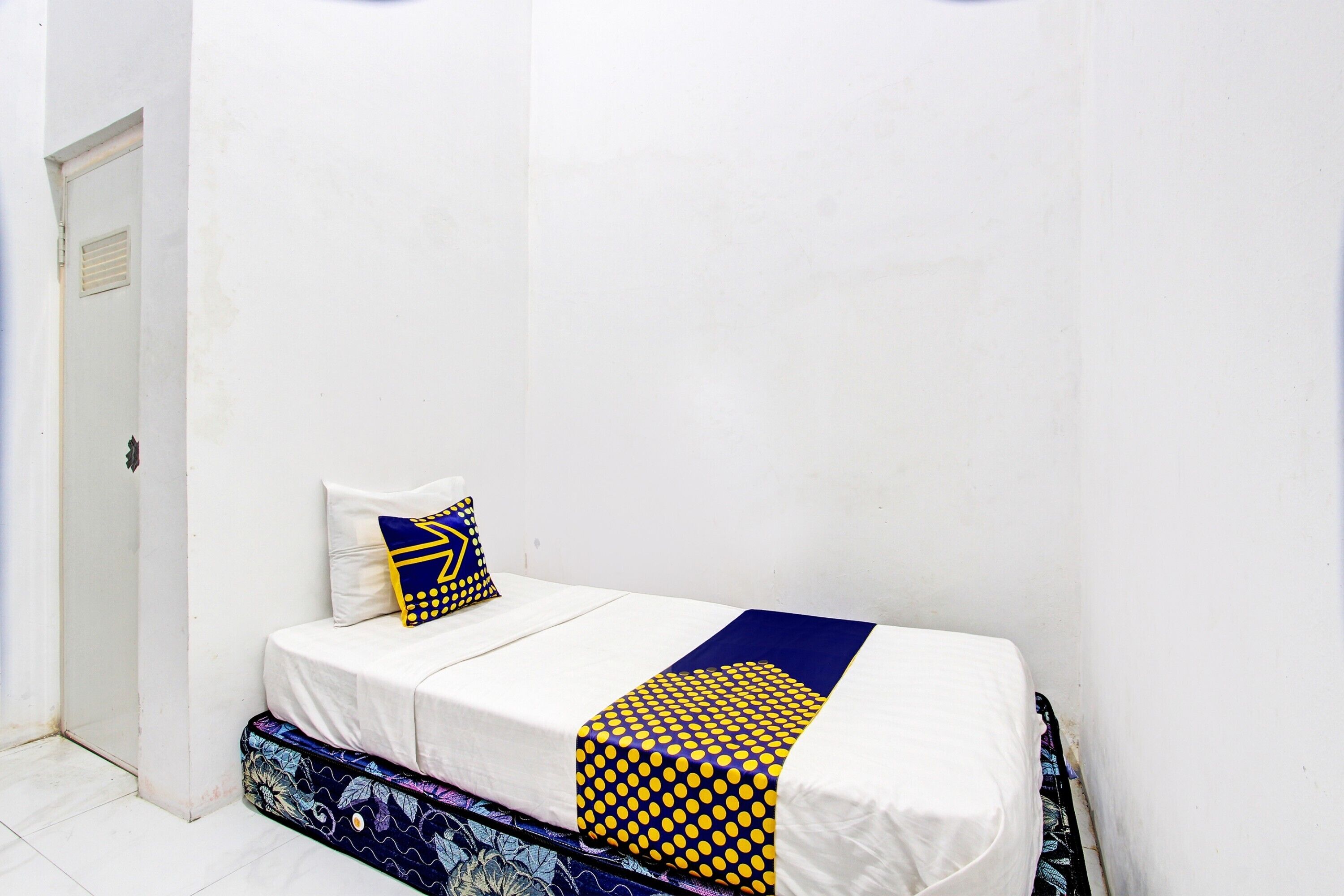 A small, single-bed room with a white bedspread, a blue and yellow patterned blanket, and a single decorative pillow.