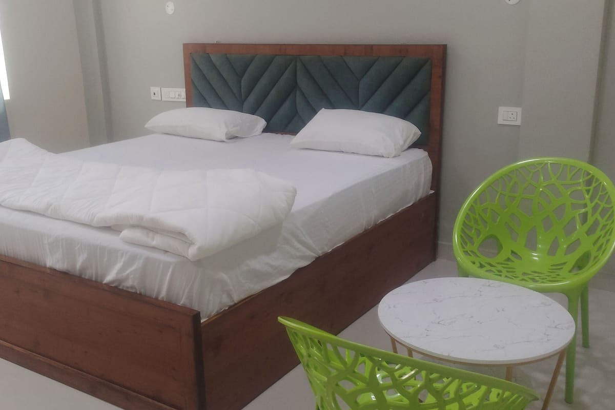 Una cama bien hecha con sábanas blancas, cabecera de madera y dos almohadas. Hay una mesita redonda con tapa de mármol blanco y una silla verde con diseño de celosía.