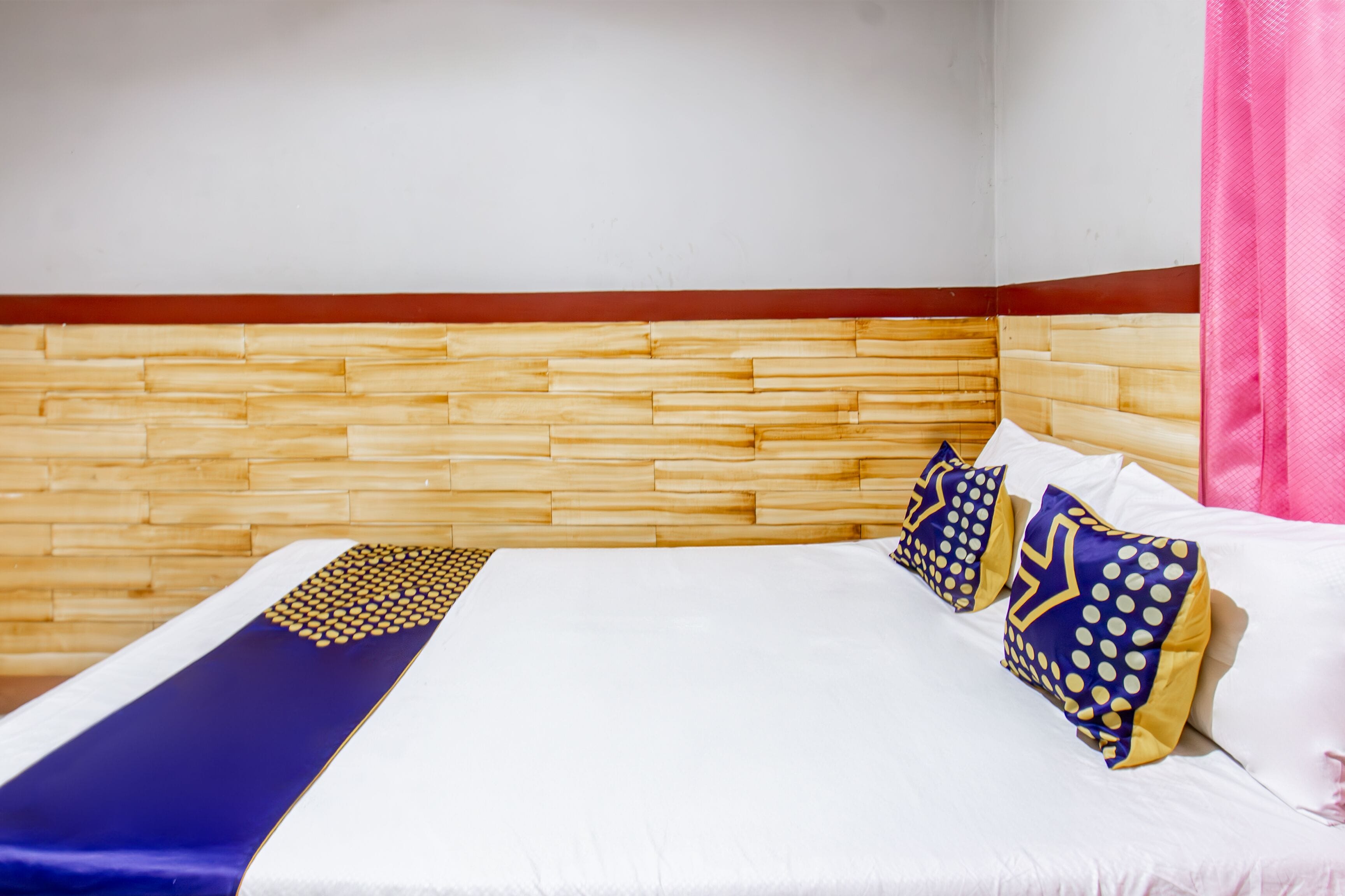 A hotel room with a wooden headboard, a white bed with blue and yellow pillows, and a pink curtain.