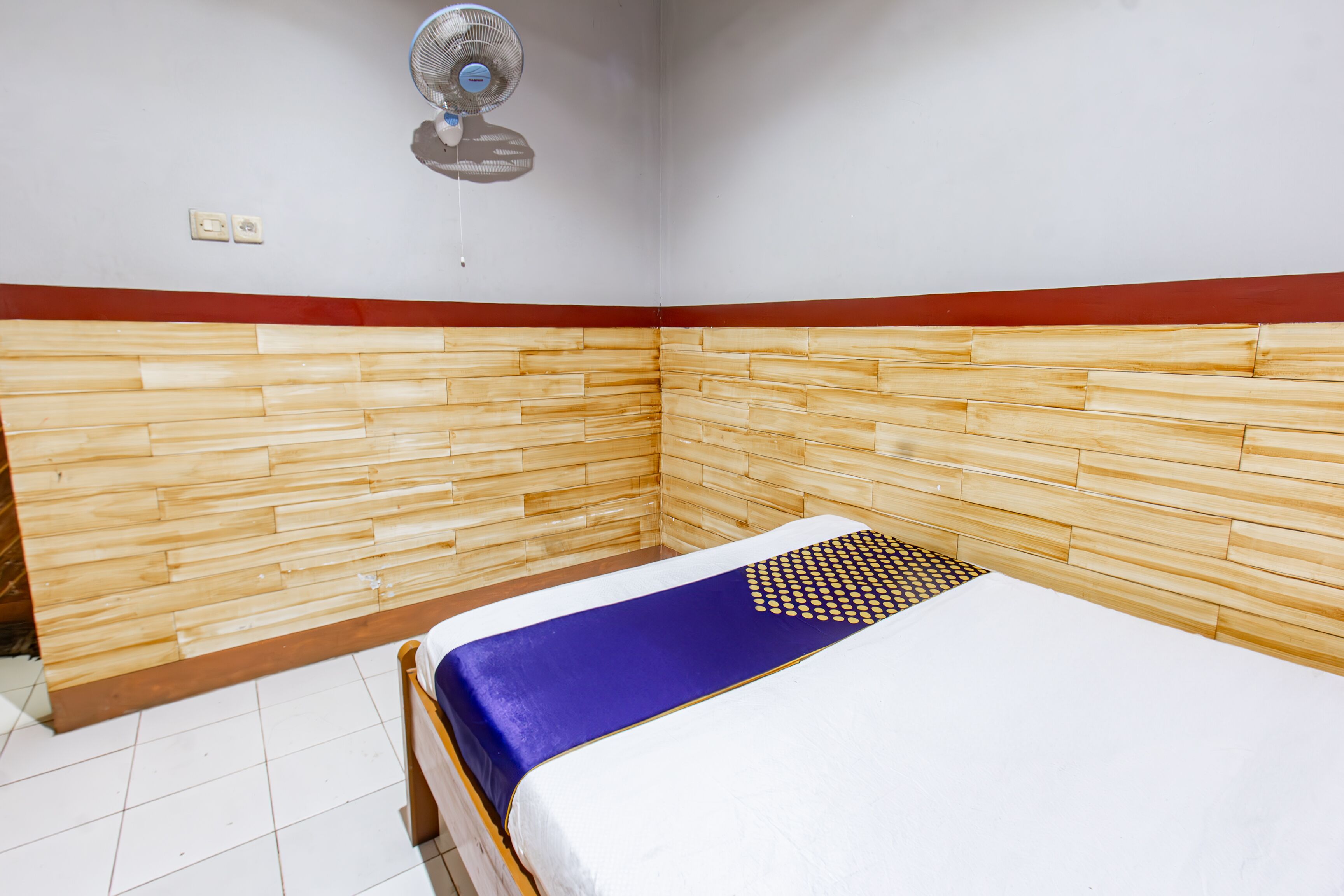 A small, neatly arranged room with a bed, a wall-mounted fan, and a red and white border.