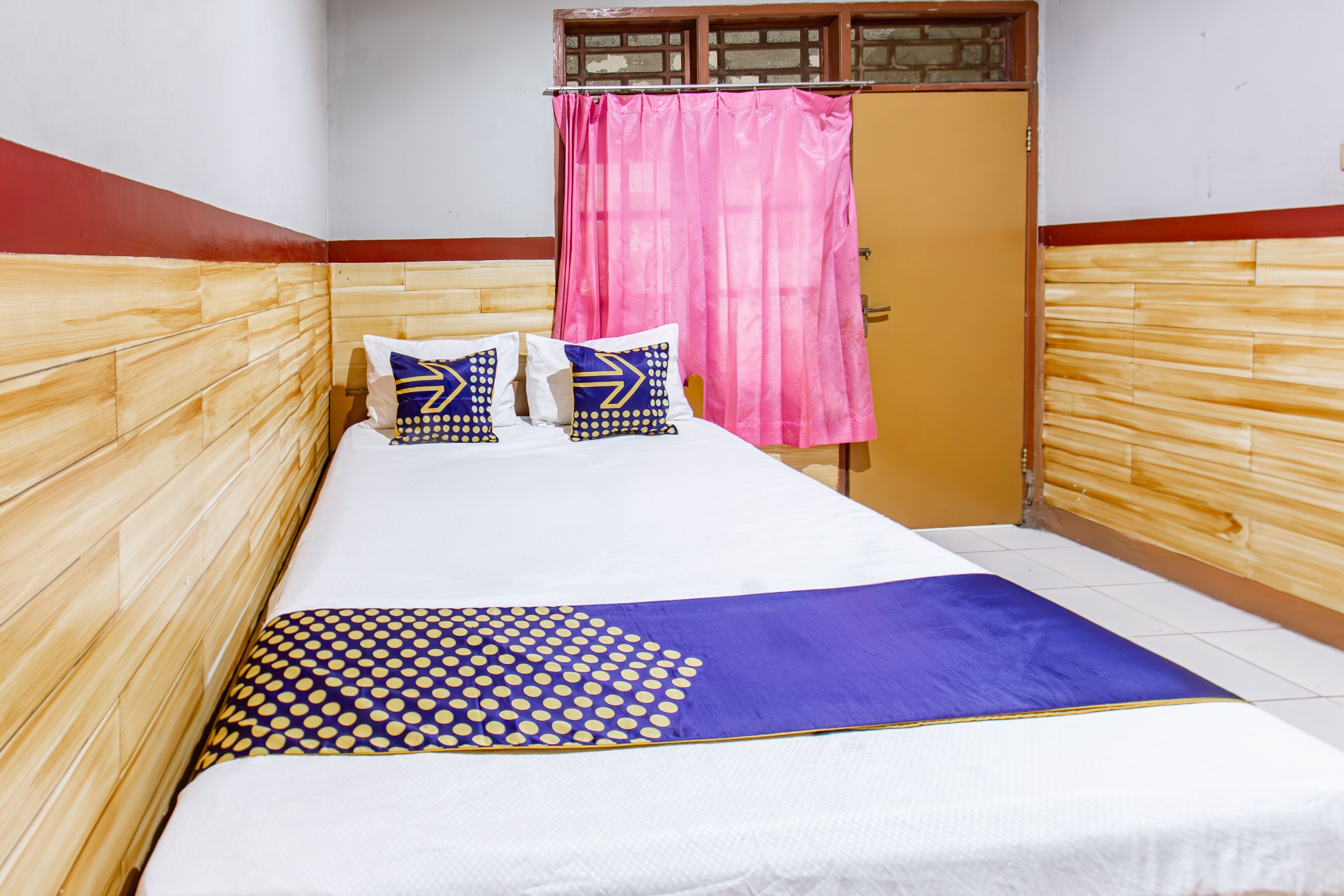 Una cama individual con colcha blanca y morada, dos almohadas con diseños azules y amarillos, y cabecera de madera.