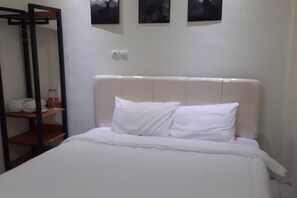 Cama bem arrumada com lençóis brancos em quarto de hotel. Há uma prateleira de madeira com toalhas e artigos de higiene pessoal.
