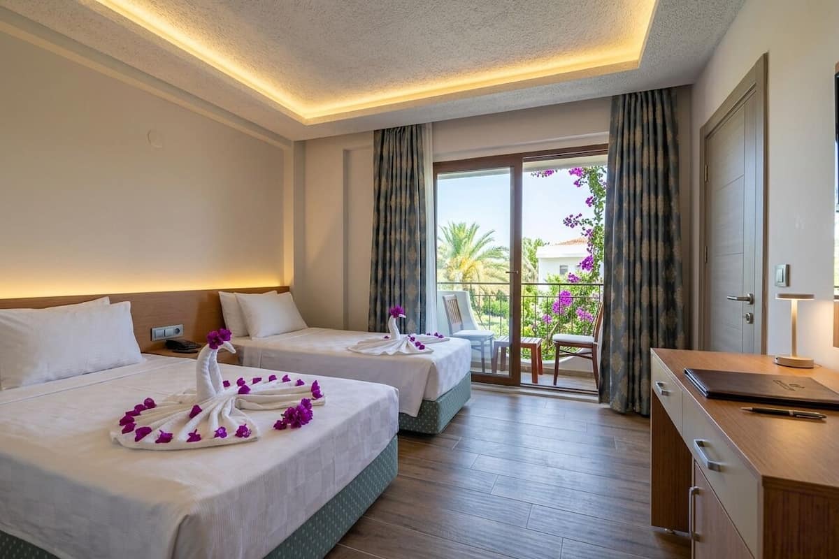 A hotel room with two beds, a desk, a chair, and a balcony with a view of palm trees and flowers.