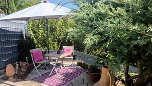 Een terras met een tafel, stoelen, een grote parasol, plantenbakken en een boom.