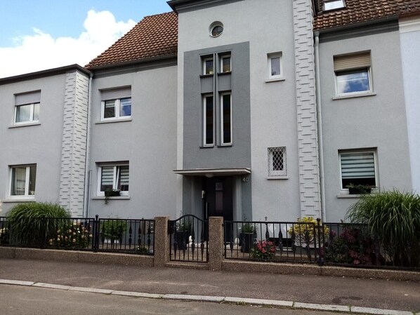 A two-story residential building with a grey facade, a tiled roof, and a gated entrance.
