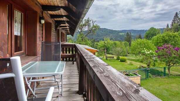 Un balcón de madera con una mesa y sillas de vidrio, con vistas a un jardín exuberante y colinas boscosas.