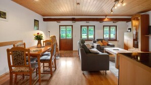 Techo de madera, mesa de comedor con sillas, sofá, zona de cocina y ventanas con cortinas.