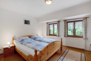 A bedroom with a wooden bed, two windows with curtains, a nightstand, a radiator, and a framed picture on the wall.