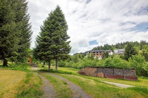 Un paysage rural avec un chemin de gravier, de grands arbres à feuilles persistantes, une petite construction en bois et, au fond, un bâtiment doté de nombreuses fenêtres.