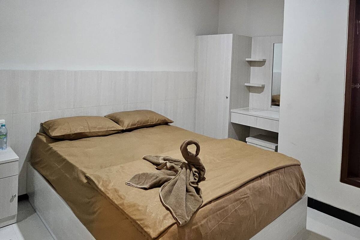 Una cama bien tendida con cubrecama y almohadas marrones, una mesita de noche blanca con una botella y un armario con espejo.