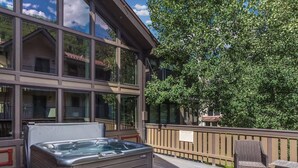 Un jacuzzi en una terraza de madera con vista a un edificio con amplios ventanales y un árbol.
