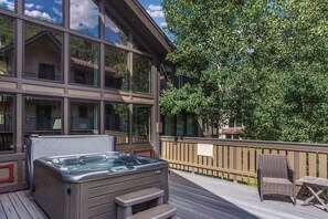 Una vasca idromassaggio su un pontile di legno, con vista su un edificio con ampie vetrate e un albero.