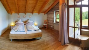 Un dormitorio con techo de madera, cama con ropa de cama floral, una silla, una mesita y una ventana con vistas a zonas verdes.