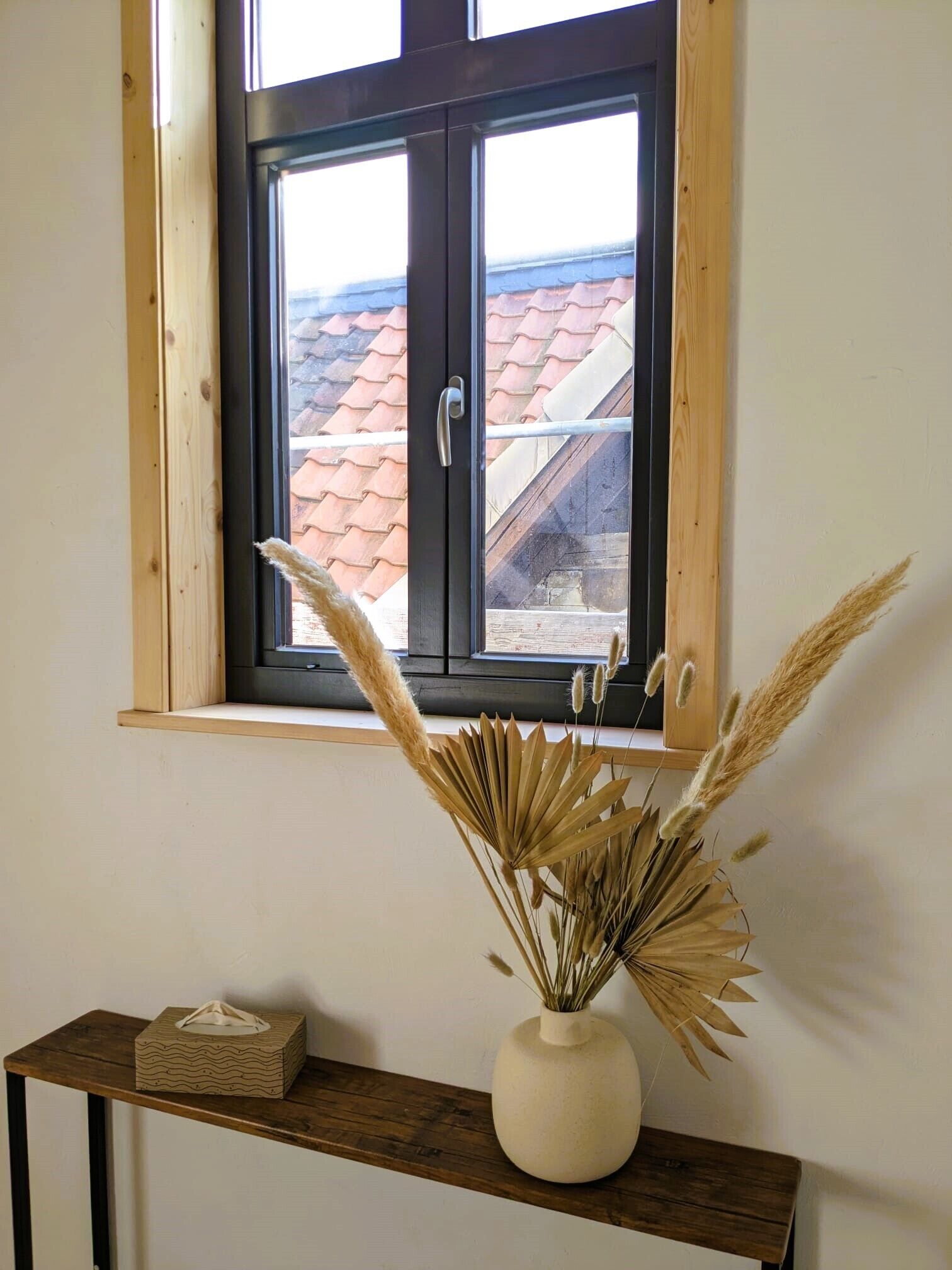 A window with a view of a red-tiled roof, a wooden shelf with a vase of dried plants, and a tissue box.
