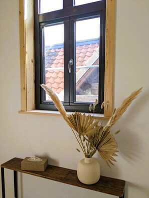 Une fenêtre donnant sur un toit recouvert de tuiles rouges, une étagère en bois avec un vase contenant des plantes séchées et une boîte de mouchoirs.