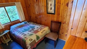 A wooden cabin bedroom with a bed, a chair, a small table, and a window.