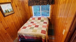 Une chambre dans une cabane en bois, avec un lit, une fenêtre donnant sur les arbres et un tableau encadré au mur.