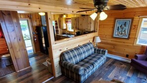 Ein Holzblockhaus-Innenraum mit einem Karo-Sofa, einem Esstisch und einer durch einen offenen Durchgang sichtbaren Küchenzeile.