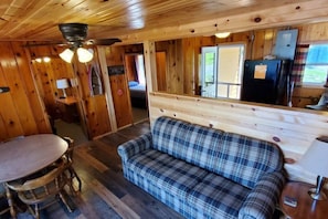 A wooden cabin interior with a plaid sofa, dining table, and ceiling fan.