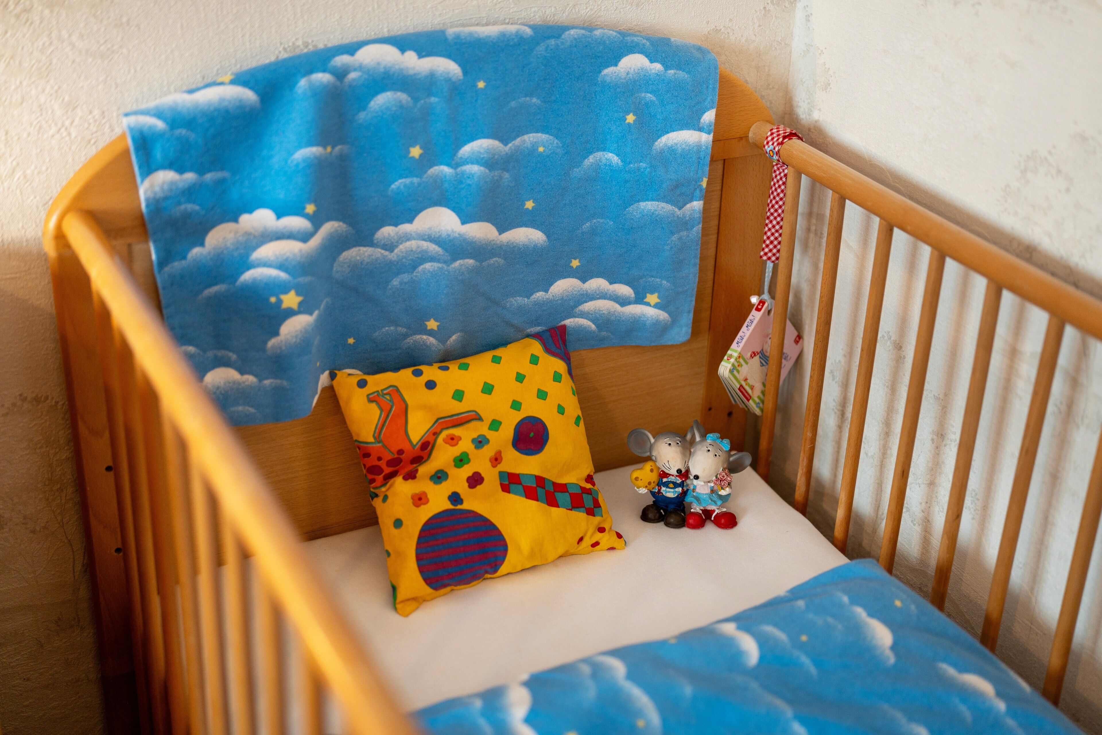A wooden crib with a blue cloud-patterned blanket, a multicolored pillow, and a small toy mouse.