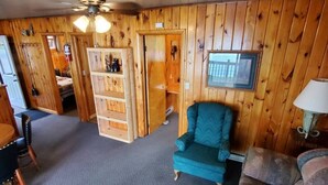 Ein Zimmer mit Holzwänden, einem türkisfarbenen Sessel, einem gemusterten Sofa, einem Esstisch mit Stühlen und einem Deckenventilator.