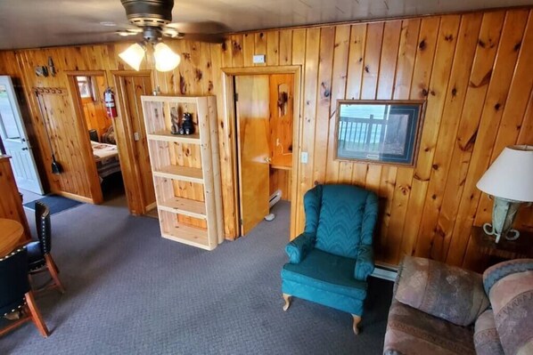 A room with wooden walls, a teal armchair, a patterned sofa, a dining table with chairs, and a ceiling fan. - Lakeside 2 Bedroom Pelican Cabin on Kab (Kabetogama)