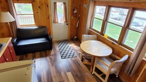 A cozy cabin interior with a wooden floor, a black leather sofa, a dining table with two chairs, and large windows offering a view of the outdoors.