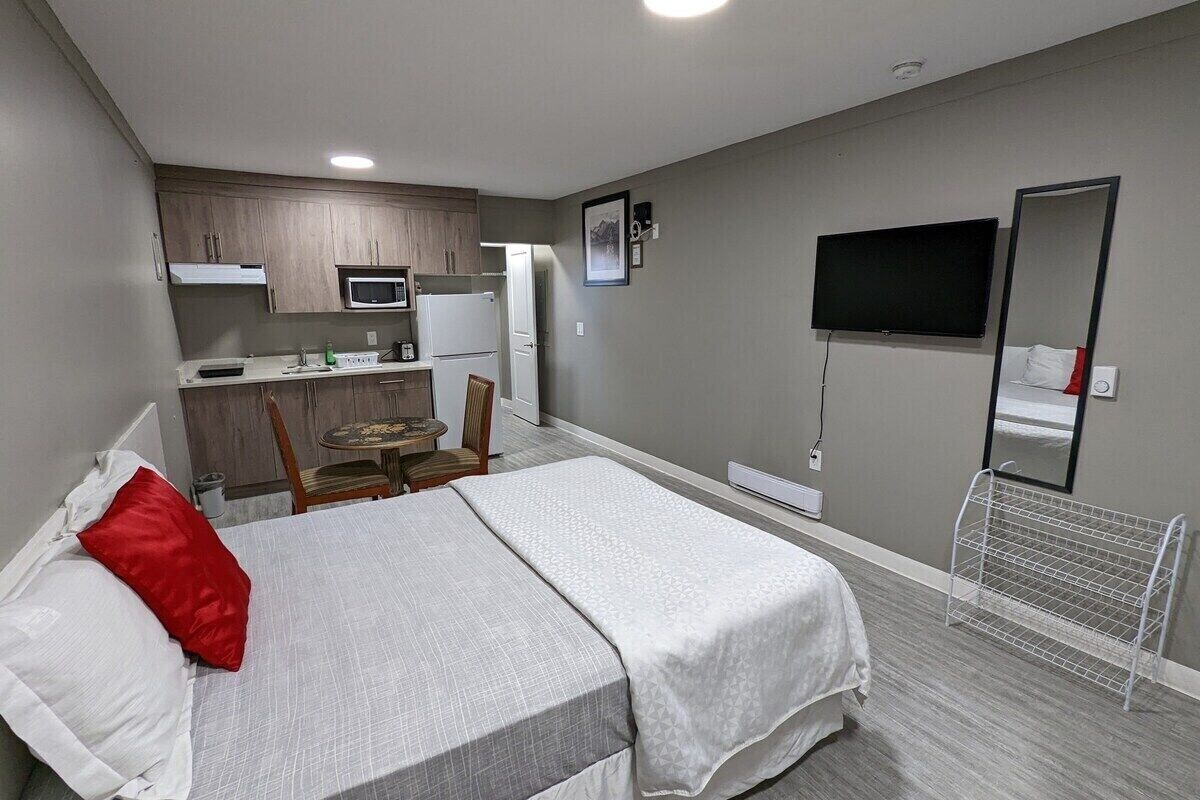 A compact hotel room with a bed, a small kitchenette, a TV, and a mirror.